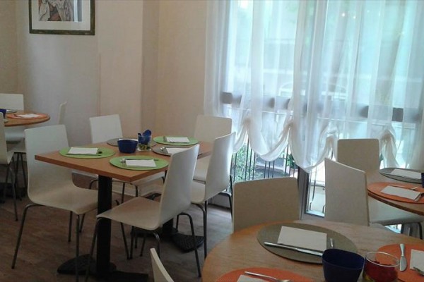 Restauracja z okrągłymi stołami, białymi krzesłami i jasnym światłem dziennym wpadającym przez zasłony.