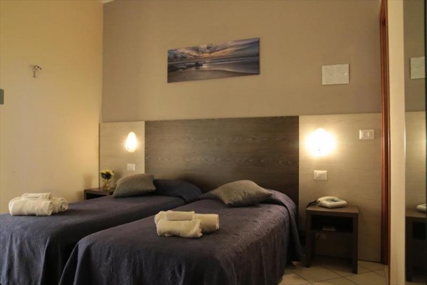 Dwa łóżka z ciemnymi narzutami w pokoju hotelowym z szafkami nocnymi i obrazem na ścianie.