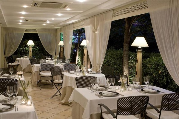 Elegancka restauracja z białymi obrusami, stylowymi krzesłami i lampami przy oknach z widokiem na ogród.