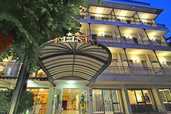 Wejście do oświetlonego czteropiętrowego hotelu z drzewami po bokach.