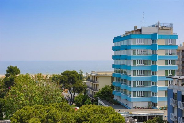 Niebiesko-biały wielopiętrowy hotel nad morzem, otoczony zielenią.