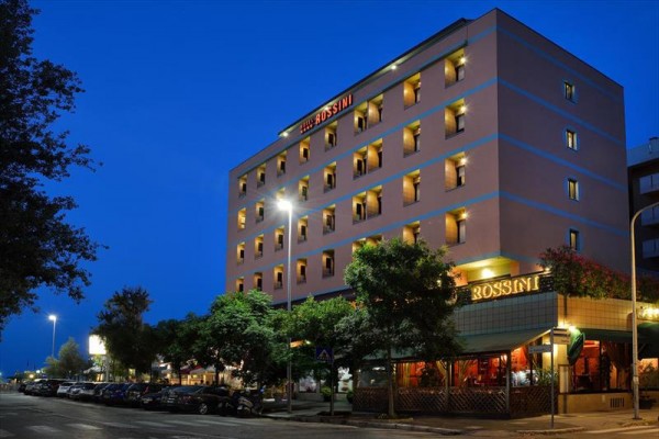 Nocny widok na oświetlony budynek hotelu Rossini otoczony drzewami.