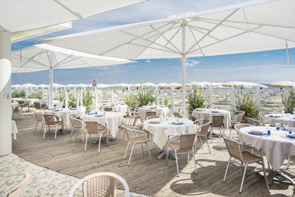 Taras restauracji z białymi stolikami i krzesłami pod parasolami z widokiem na plażę.