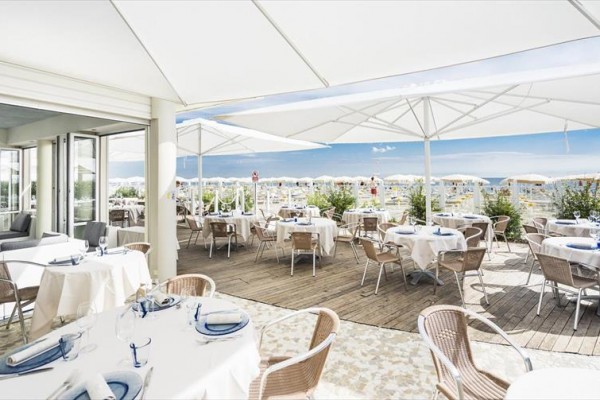 Restauracja z zewnętrznym tarasem, przykrytym białymi parasolami, z widokiem na plażę.