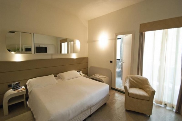 Nowoczesny pokój hotelowy z łóżkiem małżeńskim i fotelem obok okna z przeszklonymi drzwiami.