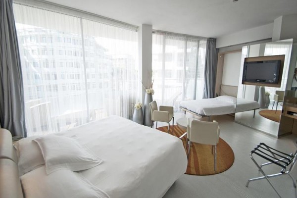 Nowoczesny pokój hotelowy z dużym łóżkiem, fotelami i oknami od podłogi do sufitu.