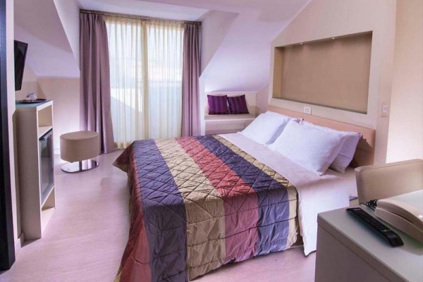 Nowoczesny pokój hotelowy z kolorową narzutą na łóżku i dużymi oknami z fioletowymi zasłonami.
