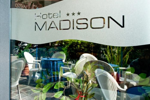 Logo hotelu Madison na szklanych drzwiach z odbiciem krzeseł i roślin.