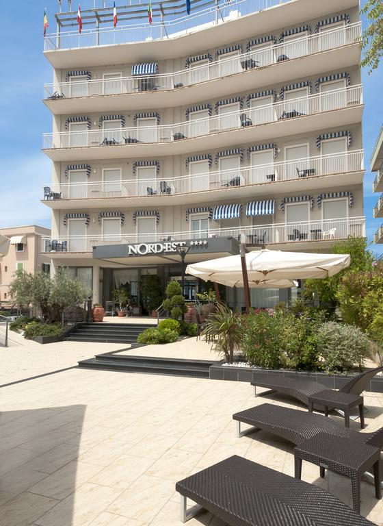 Budynek hotelu Nord Est z wieloma balkonami i zewnętrznym miejscem do siedzenia pod parasolem.