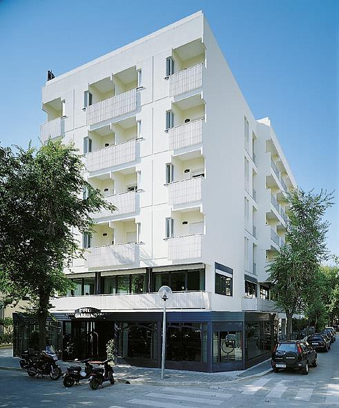 Biały budynek hotelu z balkonami na kilku piętrach pośrodku ulicy z zaparkowanymi samochodami i motocyklami.