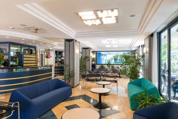 Elegancki hol hotelowy z zielonymi roślinami, niebieskimi i zielonymi sofami oraz szklanymi ścianami.