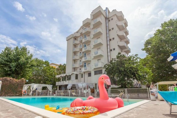 Hotelowy basen z dmuchanym różowym flamingiem i kołem przed białym budynkiem.