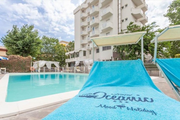 Leżak z niebieskim ręcznikiem przy hotelowym basenie przed budynkiem z balkonami otoczonym drzewami.