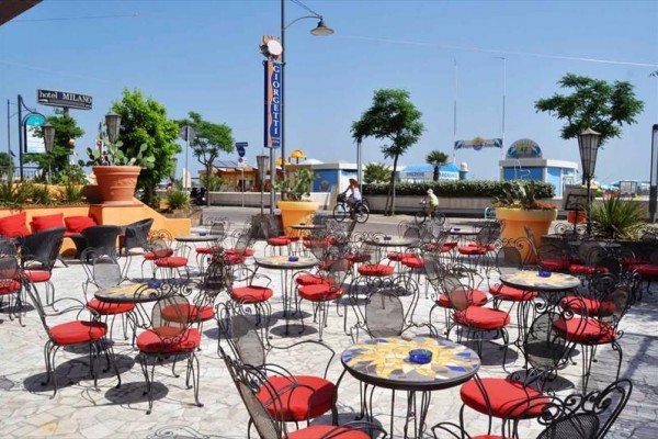 Zewnętrzna kawiarnia z metalowymi stolikami i krzesłami z czerwonymi poduszkami przy plaży.