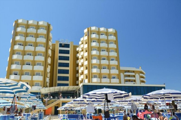 Duży hotel z balkonami i niebiesko-białymi parasolami plażowymi na piaszczystej plaży.