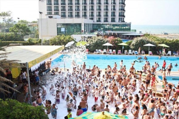 Ludzie bawią się na imprezie z pianą przy basenie przed hotelem z widokiem na plażę.