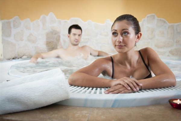 Dwie osoby relaksują się w jacuzzi otoczonym kamienną ścianą.