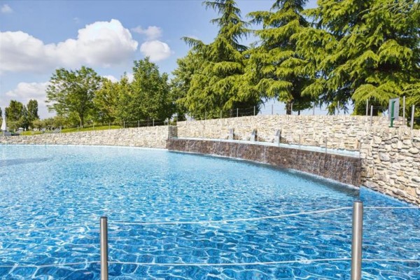 Niebieski basen z wodospadami otoczony kamiennym murem i drzewami w tle.