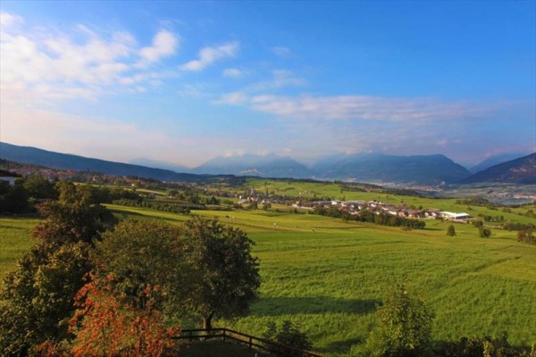 Rozległy krajobraz z łąkami, drzewami i górami w tle.