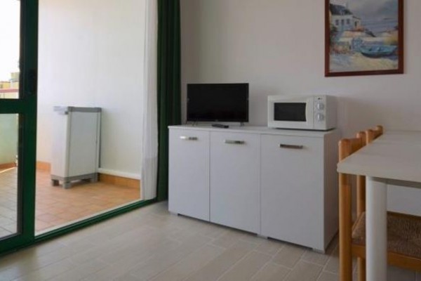 Wnętrze pokoju z białymi meblami, telewizorem i kuchenką mikrofalową, obok balkon z plastikowymi meblami.
