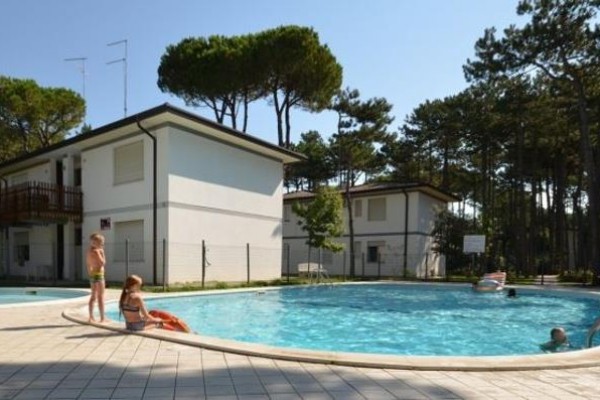 Basen wśród sosnowego lasu z kąpiącymi się ludźmi i budynkami w tle.