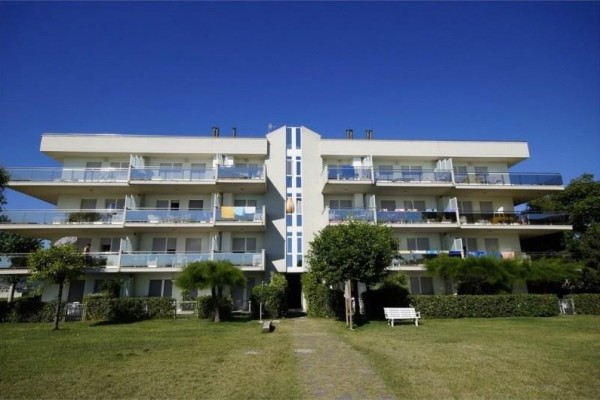 Czteropiętrowy budynek mieszkalny z balkonami i ogrodem przed nim na tle niebieskiego nieba.