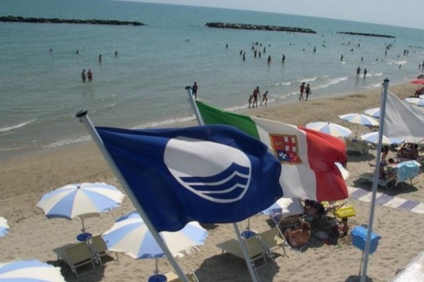 Plaża z niebieską flagą, włoską flagą i ludźmi kąpiącymi się w morzu.