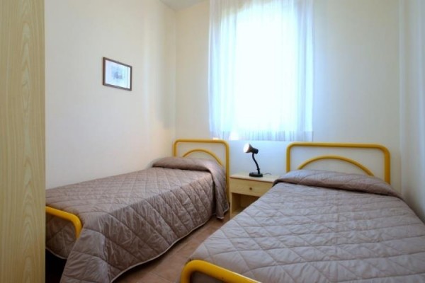 Dwa pojedyncze łóżka z beżowymi narzutami, stolik nocny i lampka pod oknem.