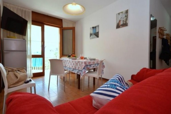 Salon z czerwoną kanapą, stołem z obrusem, krzesłami i przeszklonymi drzwiami na balkon.