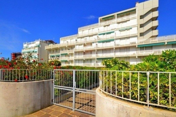 Blok mieszkalny z białymi balkonami otoczony zielenią pod niebieskim niebem.