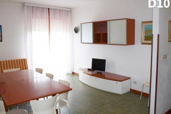 Prosto urządzony salon z telewizorem, stołem i krzesłami przy dużym oknie z białymi zasłonami.