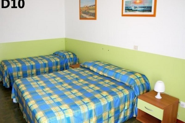 Dwa jednoosobowe łóżka z niebiesko-żółtymi kraciastymi narzutami i stolikiem nocnym z lampką w pokoju.