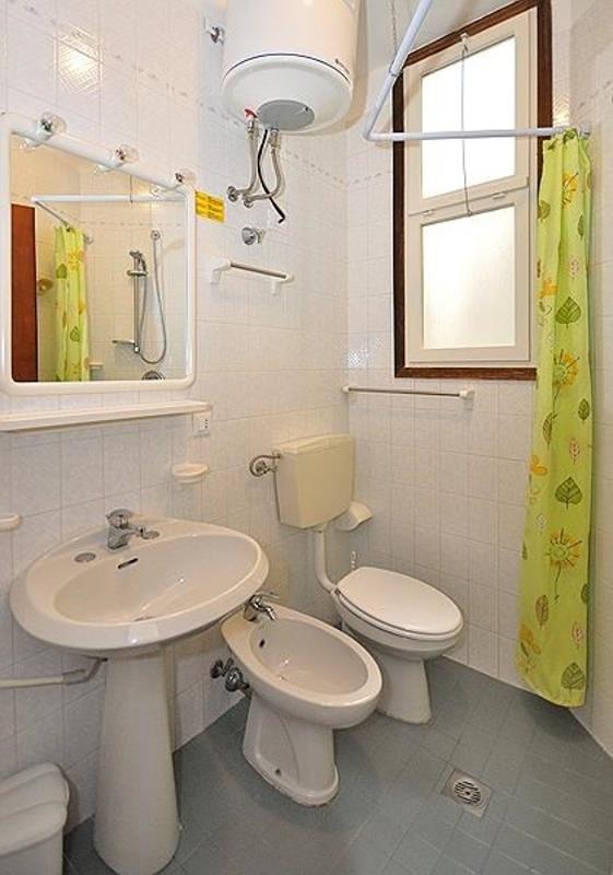 Łazienka z ceramiczną umywalką, bidetem i toaletą, prysznic za zieloną zasłoną i podgrzewacz wody na ścianie.