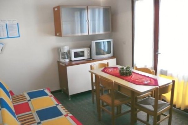 Prosto urządzony salon z kanapą, stołem, krzesłami, telewizorem i kuchenką mikrofalową.